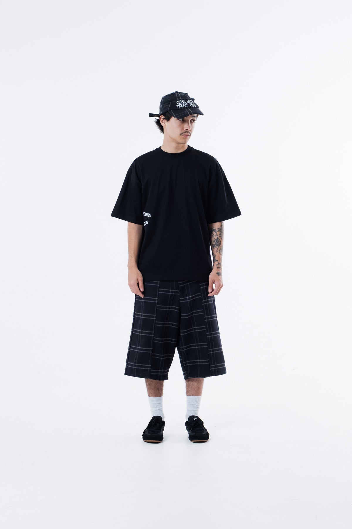 Modelo veste a Freak Show Classic Tee preta com bone e bermuda xadrez, vista frontal de corpo inteiro