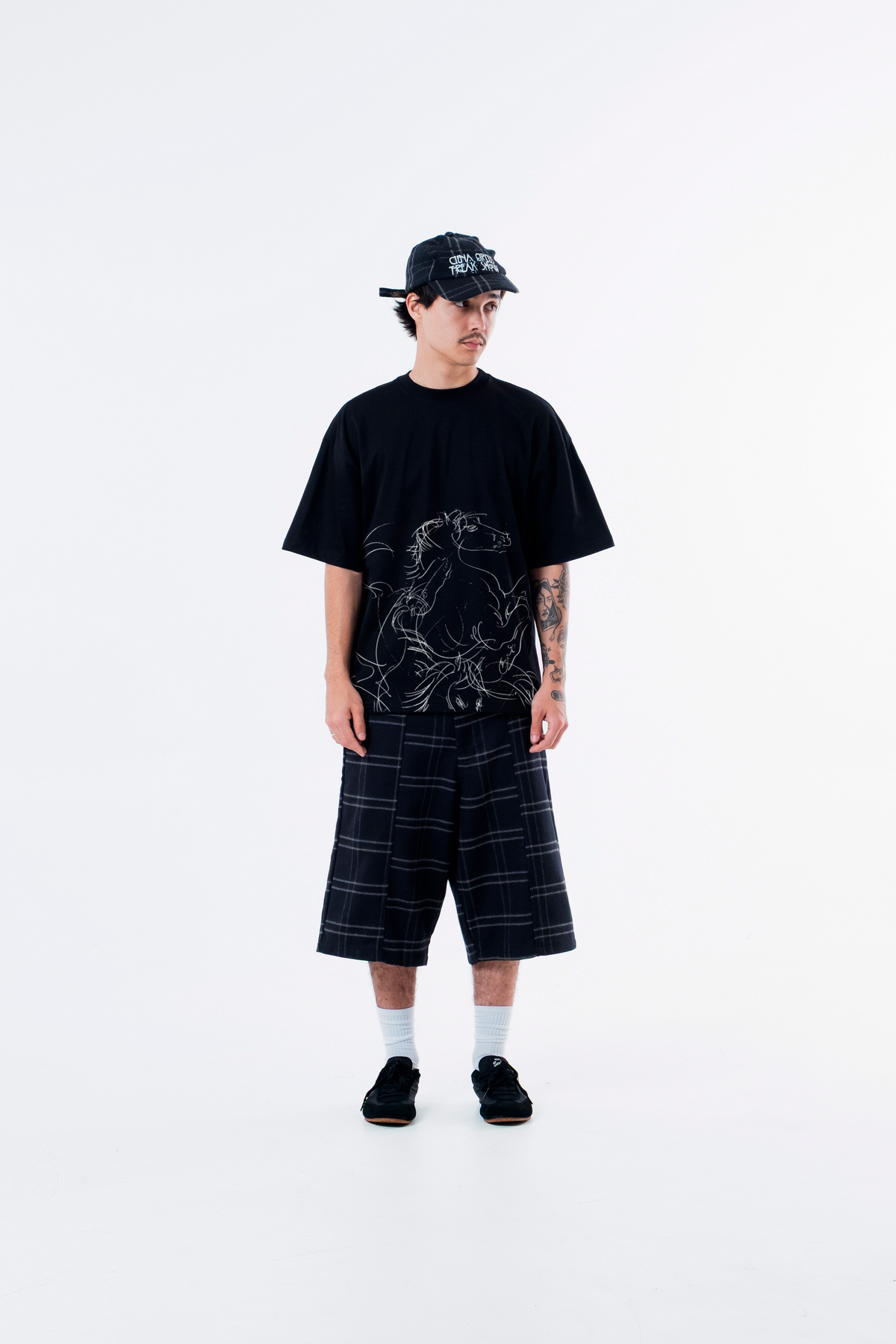 Modelo veste a Cheval Black Heavy Tee preta com bone e bermuda xadrez, vista frontal de corpo inteiro